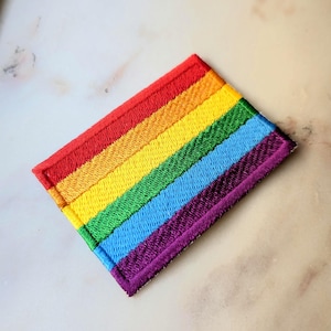 Può includere: Una toppa rettangolare ricamata con un vivace design arcobaleno a strisce orizzontali nei colori rosso, arancione, giallo, verde, blu e viola. I bordi della toppa sono rifiniti con un bordo in filo denso coordinato per un aspetto pulito.