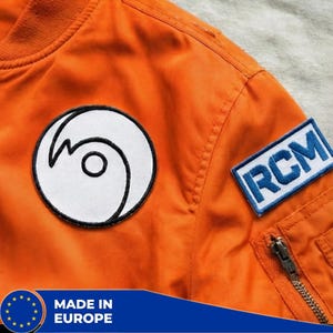Op de afbeelding: Close-up van een oranje bomberjack met een wit en zwart cirkelvormig embleem en een rechthoekig embleem met "RCM" in blauw. De jas heeft een rits en is gemaakt in Europa.