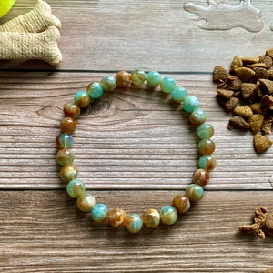 May include: A beaded bracelet with brown and teal-colored beads. The bracelet is on a wooden surface with dog treats, a tennis ball, and a dog bowl. The beads are round and have a marbled appearance.