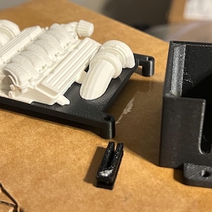 May include: A collection of 3D-printed model car engine parts. The engine block and components are white, mounted on a black base. A black rectangular holder and a small black piece are also present.