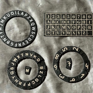 May include: A set of black glittery items including a circular ring with letters, a rectangular grid with numbers 1-9 and letters A-Z, and two circular rings featuring individual letter and number tiles, with one letter M and one number 7 tile detached.