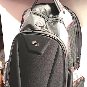 May include: Black backpack with red accents, hanging from a black hook. The backpack features a padded front panel with a silver logo, a zippered front pocket, and mesh side pockets. The backpack is made of a durable, textured material.