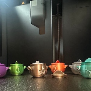 May include: Six small, 3D printed ceramic pots with lids. The pots are in various colors, including purple, green, gold, orange, silver, and teal. Each pot has a different design on the lid, including a crescent moon, a star, and a pentagram.