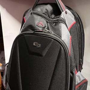 May include: Black backpack with red accents and a gray mesh panel. The backpack has a large front pocket with a silver logo that says "G".