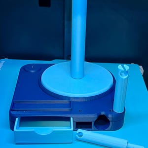 May include: A blue 3D-printed filament spool holder with a drawer. The holder has a dark blue base, a light blue disc, and a tall, cylindrical light blue post. A smaller light blue piece is next to the holder.