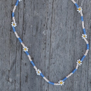 May include: A blue and white beaded necklace with small white flowers and yellow centers. The necklace has a gold clasp.