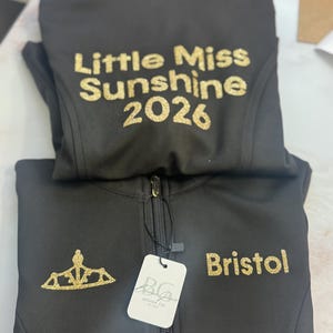 May include: Black zip-up jacket with gold glitter text that reads "Little Miss Sunshine 2026" on the back. The front features a gold glitter crown and the name "Bristol". A tag from "Boheme Grace Design Co." is attached.