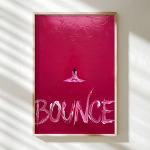 May include: A framed artwork with a textured, deep pink background. A small figure in a pink dress is centered, above the word "BOUNCE" in white, brushstroke-style lettering. The artwork is in a light wood frame.