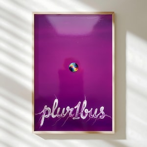 May include: A framed art print with a vibrant purple background. A small, colorful sphere is centered, with the word "plur1bus" written in white script below. The artwork is encased in a natural wood frame.