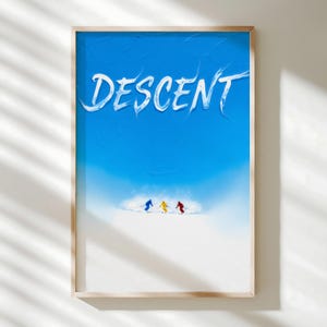 Op de afbeelding: Een ingelijste print met een blauwe lucht en het woord "DESCENT" in witte letters. Drie skiërs in blauw, geel en rood skiën een besneeuwde helling af. Het kunstwerk zit in een houten lijst.