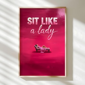 May include: A framed art print with a vibrant pink background and the text "SIT LIKE a lady" in white script. The image features a woman in a pink dress reclining on a chaise lounge, creating a stylish and elegant aesthetic.