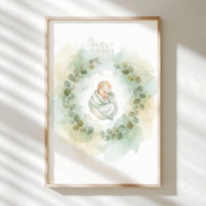 Könnte beinhalten: Ein gerahmter Aquarelldruck mit einem schlafenden Baby, das in eine Decke gewickelt ist und von einem Kranz aus grünen Blättern umgeben ist. Die Worte "SLEEP GRACE" sind in Gold über dem Baby geschrieben. Der Hintergrund hat weiche Aquarellwaschungen in Grün- und Gelbtönen.