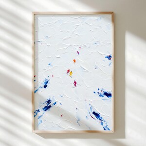 May include: Abstract art print of skiers on a snowy mountain. The artwork features a textured white background with blue and red accents. Small figures in bright colors, including yellow, orange, and pink, are skiing down the mountain. The print is framed in a light wood.