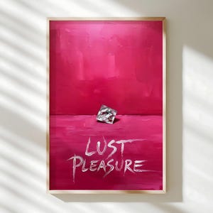 May include: A framed artwork with a vibrant pink background and a silver object. The words "Lust Pleasure" are written in white paint across the bottom. The artwork is encased in a light brown wooden frame.