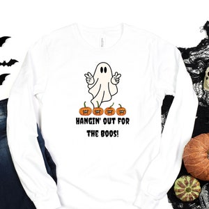 May include: White long-sleeved t-shirt with a ghost wearing a peace sign and the text "Hangin' out for the boos!"  Four orange pumpkins with spider accents are below the ghost.