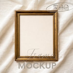 May include: A rectangular gold-colored frame with a cream-colored interior, resting on a white fabric surface. The word "Frame" is written in cursive script, and the word "MOCKUP" is below it in a sans-serif font.