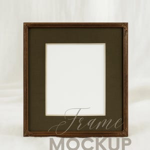 May include: A square brown picture frame with a dark green mount and a white interior. The frame has ornate detailing. The word "Frame" is written in cursive, and "Mockup" is below it. The background is white.