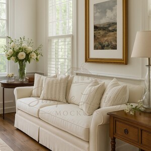 May include: A cream sofa with striped cushions and a framed landscape painting. The room includes a wooden side table, a lamp, and a vase of white flowers. The decor creates a comfortable and elegant atmosphere.