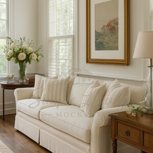 May include: A cream sofa with striped and plain cushions in a bright room. A framed picture hangs above the sofa. A side table with a lamp and a vase of flowers are also visible. The room has wooden floors and large windows.
