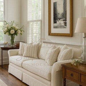May include: A cream sofa with striped and patterned cushions in a bright room with a framed painting and a table lamp. The room has wooden flooring and large windows with white shutters. A vase of white flowers sits on a small side table.