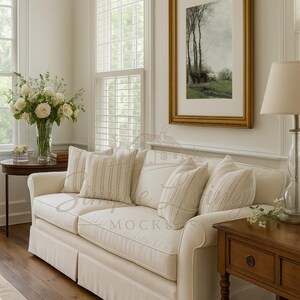 May include: A cream sofa with striped and textured cushions. A framed landscape painting hangs above the sofa. A side table with a lamp and a vase of flowers are also visible. The room has wooden flooring and large windows.