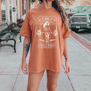 May include: A rust-coloured t-shirt with a white graphic of a woman lifting weights. The graphic is in a vintage style with the text "Muscle Mommy" below it.