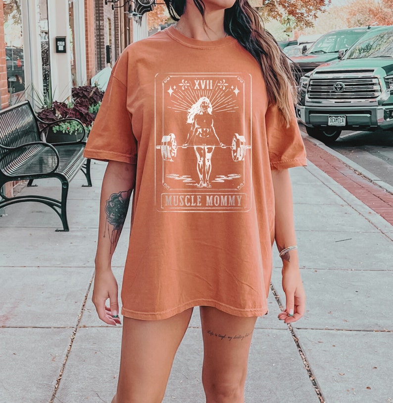 May include: A rust-coloured t-shirt with a white graphic of a woman lifting weights. The graphic is in a vintage style with the text "Muscle Mommy" below it.