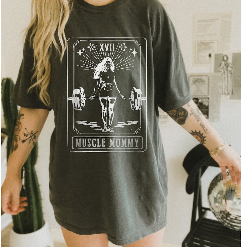 May include: A black t-shirt featuring a white graphic of a woman lifting weights. The graphic is in the style of a tarot card and reads "Muscle Mommy" at the bottom.