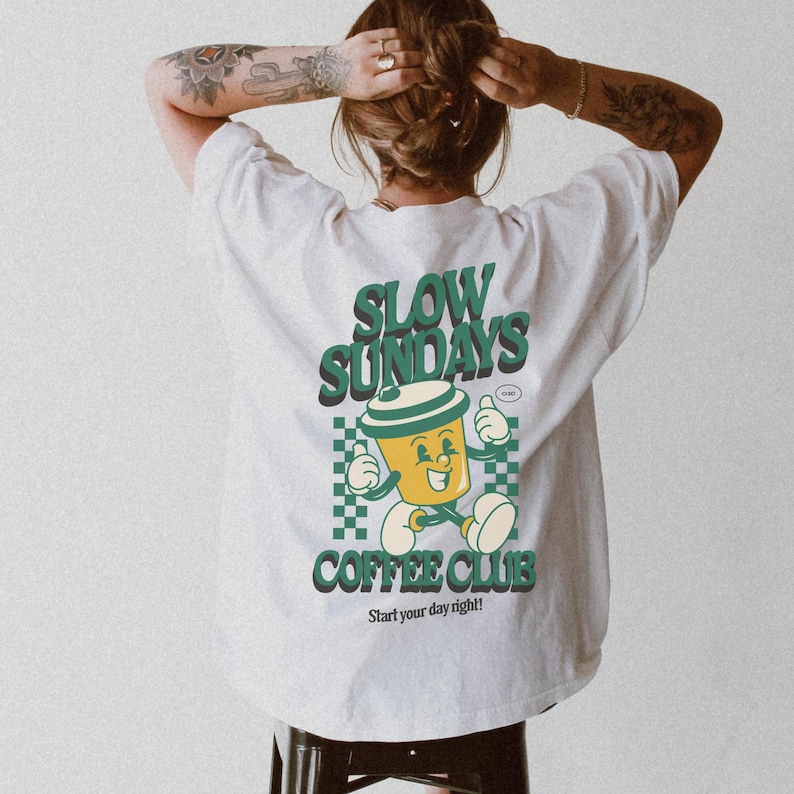 May include: Cream-colored t-shirt with a graphic on the back. The graphic features a cartoon coffee cup character with the text "SLOW SUNDAYS COFFEE CLUB" in green and white. The phrase "Start your day right!" is also included.