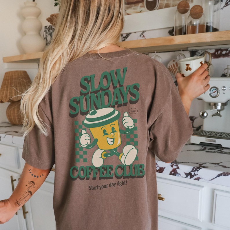 May include: Brown t-shirt with a graphic of a smiling coffee cup character and the words "SLOW SUNDAYS COFFEE CLUB". The design is in green and yellow, with the phrase "Start your day right!" at the bottom.