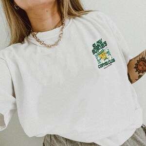 May include: White t-shirt with a green and yellow graphic that reads "Slow Sundays Coffee Club" with a cartoon coffee cup design. The shirt has a relaxed fit and is made of a soft material. A gold chain necklace is visible.
