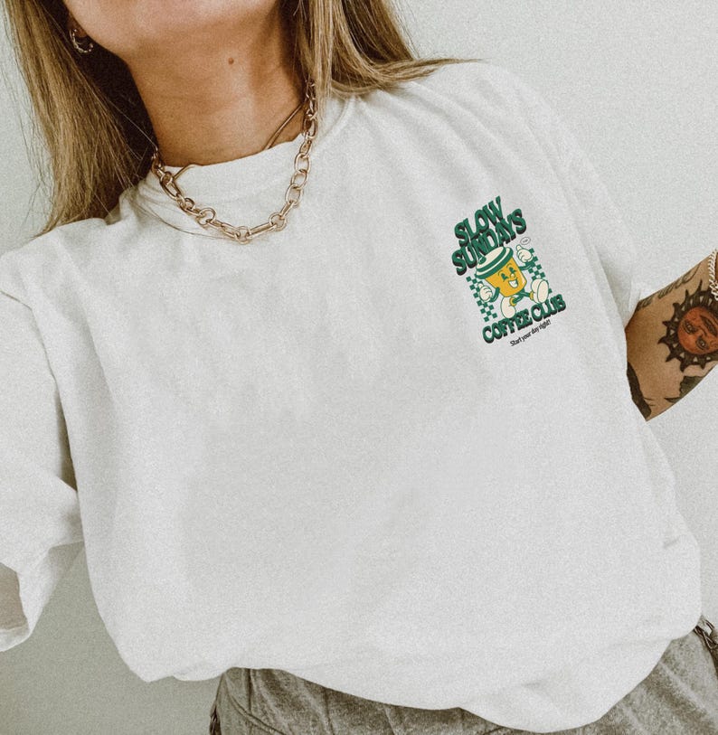 May include: White t-shirt with a green and yellow graphic that reads "Slow Sundays Coffee Club" with a cartoon coffee cup design. The shirt has a relaxed fit and is made of a soft material. A gold chain necklace is visible.