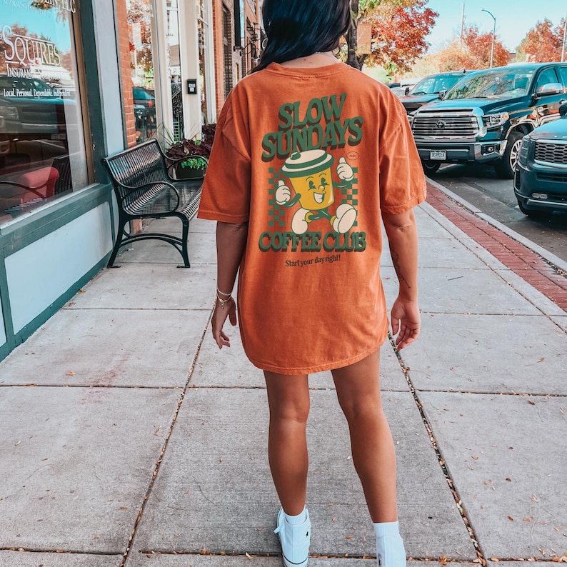 May include: An orange t-shirt with the text "SLOW SUNDAYS COFFEE CLUB" in green. The graphic features a cartoon coffee cup with arms and legs. The shirt is worn by a person standing on a sidewalk.
