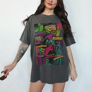May include: Charcoal grey t-shirt displaying a vibrant collage of vintage monster movie posters. The design showcases images of Frankenstein, Dracula, the Mummy, and the Wolfman, with text including "Monster," "Mummy," and "Frankenstein."