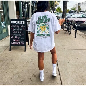 May include: White t-shirt with a green and yellow graphic that reads "SLOW SUNDAYS COFFEE CLUB" with a cartoon coffee cup. The shirt is paired with white sneakers and socks. A sign for cookies is in the background.