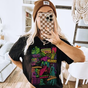 May include: Black t-shirt with a colourful graphic of classic movie monsters, including Frankenstein and Dracula. The shirt has the text "MONSTERROR" and "WOLF MAN". The person is wearing a brown beanie.