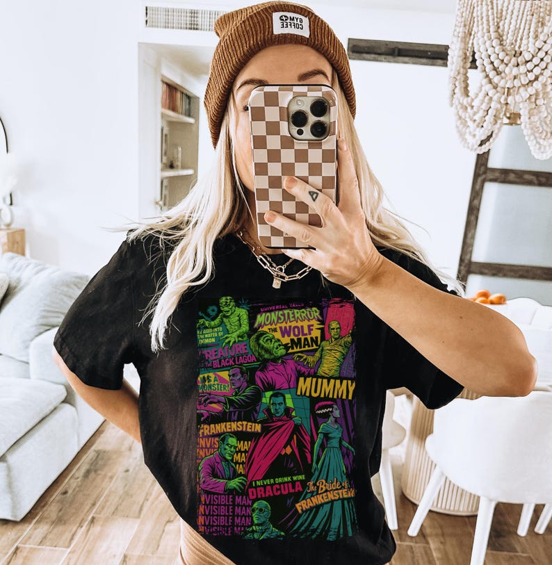 May include: Black t-shirt with a colourful graphic of classic movie monsters, including Frankenstein and Dracula. The shirt has the text "MONSTERROR" and "WOLF MAN". The person is wearing a brown beanie.