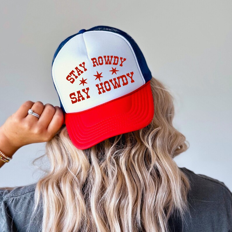 May include: A white and red trucker hat with a blue mesh back. The hat has the text "Stay Rowdy Say Howdy" in red with two stars on either side of the text.