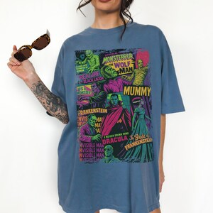 May include: A blue t-shirt with a vibrant graphic of classic Universal Monsters film posters. The design includes images of the Mummy, Frankenstein, Dracula, and the Wolfman. The t-shirt has a relaxed fit.