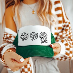 May include: A white and green trucker hat with a white front panel featuring three black and white tiger head illustrations. The text "Raisin Hell" is printed in black below the tiger heads.