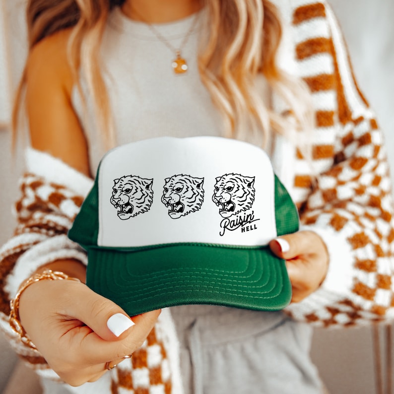 May include: A white and green trucker hat with a white front panel featuring three black and white tiger head illustrations. The text "Raisin Hell" is printed in black below the tiger heads.