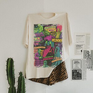 May include: A cream-coloured t-shirt with a vibrant graphic of classic movie monsters, such as Frankenstein and Dracula. The shirt is paired with animal print shorts. A cactus is in the background.