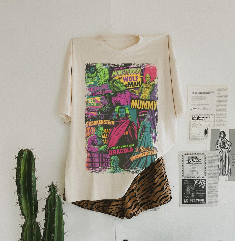 May include: A cream-coloured t-shirt with a vibrant graphic of classic movie monsters, such as Frankenstein and Dracula. The shirt is paired with animal print shorts. A cactus is in the background.