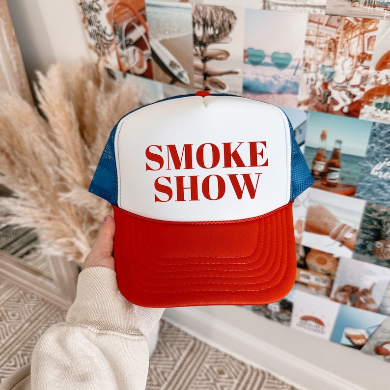 May include: A red, white, and blue trucker hat with the words "SMOKE SHOW" printed in red. The hat features a red peak, a white front panel, and a blue mesh back. A classic style of hat.