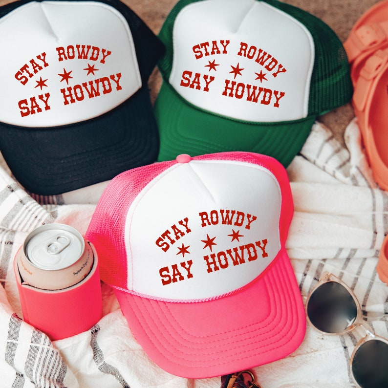 May include: Three trucker hats with a white mesh back and a white front panel. The front panel of each hat has the text "STAY ROWDY * * SAY HOWDY" in red lettering. The hats are in black, green, and pink.