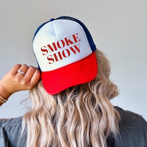 May include: A trucker hat with a white front panel, a red peak, and a navy blue mesh back. The words "SMOKE SHOW" are printed in red across the front. The hat is worn by a person with blonde hair.