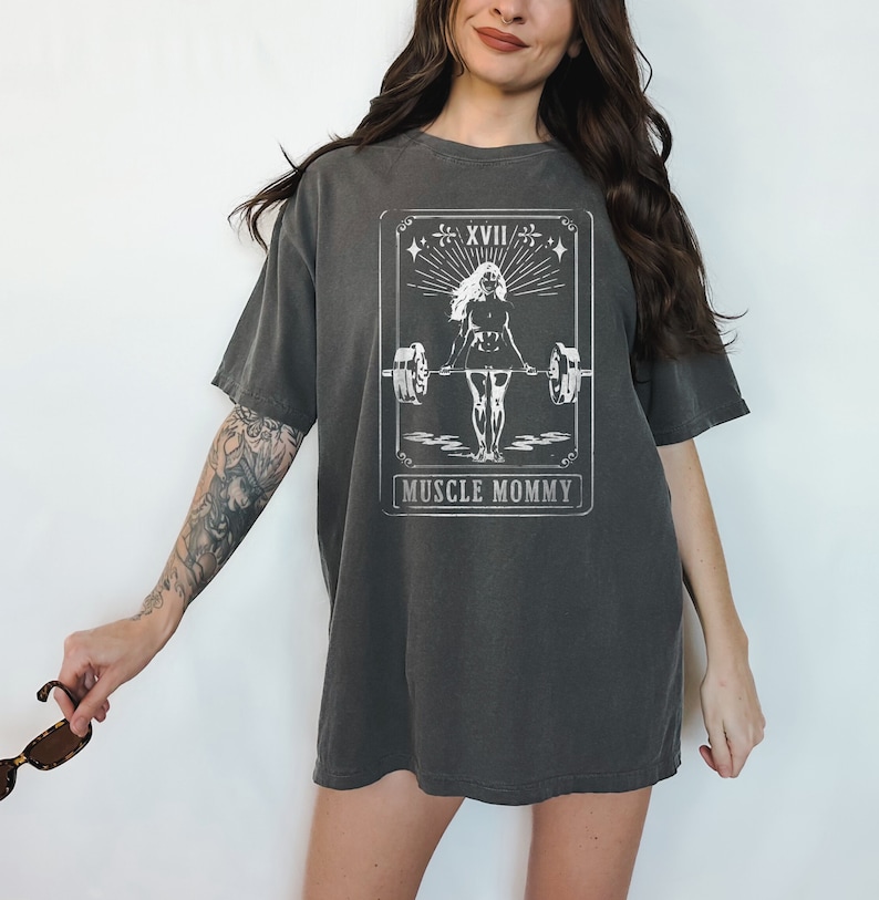 May include: An oversized t-shirt in a dark grey colour with a white graphic of a woman lifting weights. The graphic is in the style of a tarot card and says "Muscle Mommy" at the bottom.