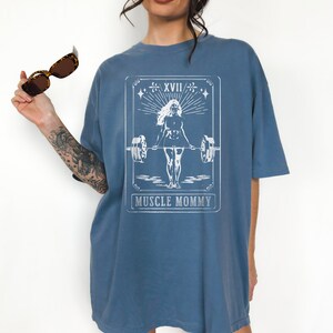 May include: An oversized blue t-shirt featuring a white graphic of a woman lifting weights. The graphic is in the style of a tarot card and reads "Muscle Mommy" at the bottom.