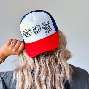 May include: A white, red, and blue trucker hat with a black and white tiger graphic and the text "raisin hell".