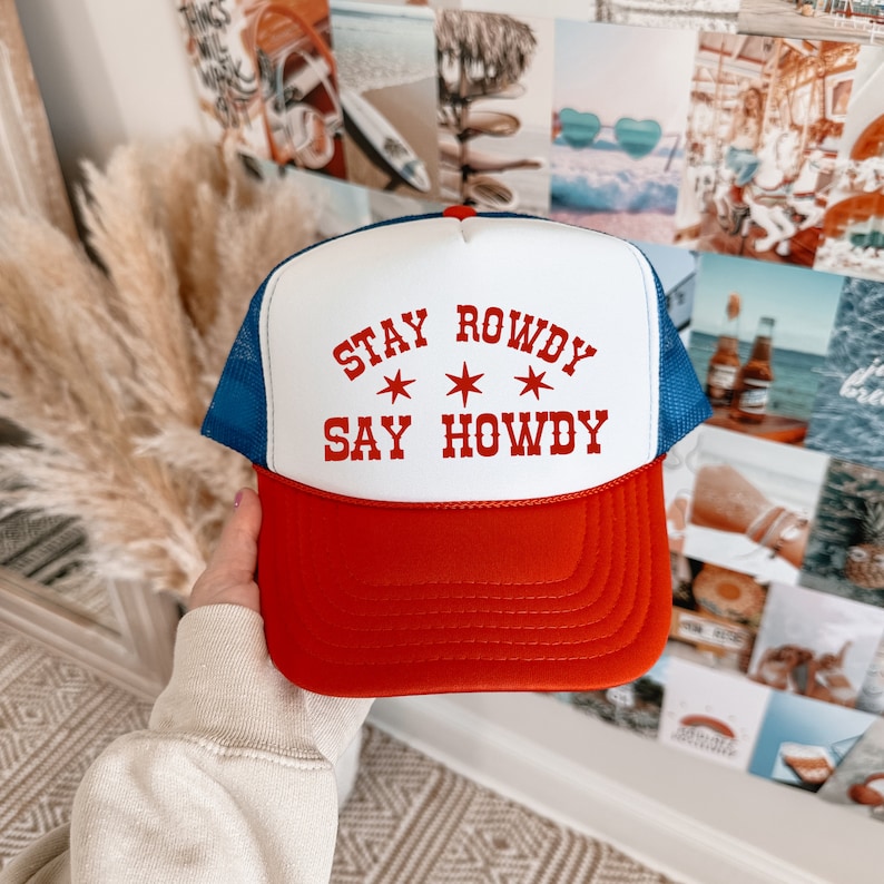 May include: A white and red trucker hat with a blue mesh back. The hat has the text "Stay Rowdy Say Howdy" in red with three white stars.